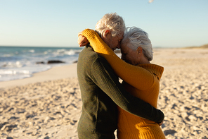 Side view of a senior Caucasian couple at the beach wearing sweaters, embracing and touching heads, with blue sky and sea in the background
