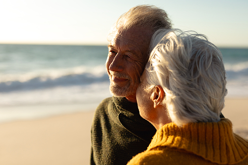 Side view of a senior Caucasian couple at the beach wearing sweaters, the woman kissing the man and the man smiling, with blue sky and sea in the background