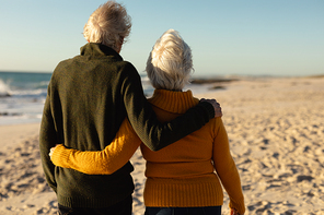 Rear view of a senior Caucasian couple at the beach wearing sweaters, embracing and walking, with blue sky and sea in the background