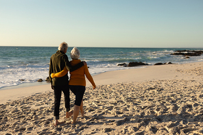 Rear view of a senior Caucasian couple at the beach wearing sweaters, embracing and walking barefoot, with blue sky and sea in the background