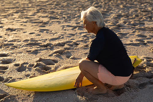 Side view of a senior Caucasian woman at the beach at sunset, kneeling down on the sand and holding a surfboard