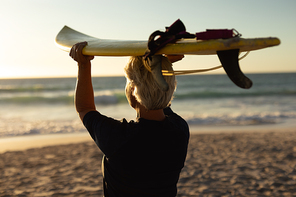 Rear view of a senior Caucasian woman at the beach at sunset, holding a surfboard on her head and looking out to sea