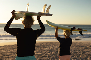 Rear view of a senior Caucasian couple at the beach at sunset, holding surfboards on their heads and looking out to sea