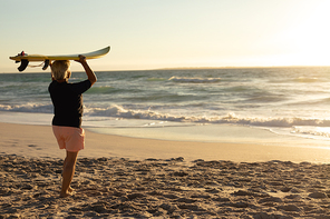 Rear view of a senior Caucasian woman at the beach at sunset, holding a surfboard on her head and looking out to sea