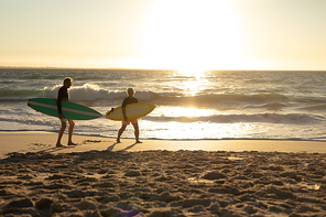 Side view of a senior Caucasian couple at the beach at sunset, holding surfboards under their arms and walking along the beach