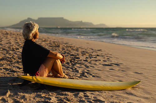 Side view of a senior Caucasian woman at the beach at sunset, sitting on the sand with a surfboard beside her, looking out to sea