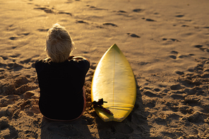 Rear view of a senior Caucasian woman at the beach at sunset, sitting on the sand with a surfboard beside her, looking out to sea