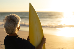 Rear view of a senior Caucasian woman at the beach at sunset, standing on the sand holding a surfboard and looking out to sea