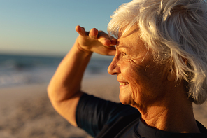 Side view close up of a senior Caucasian woman at the beach at sunset, shielding her eyes from the sun, smiling and looking out to sea
