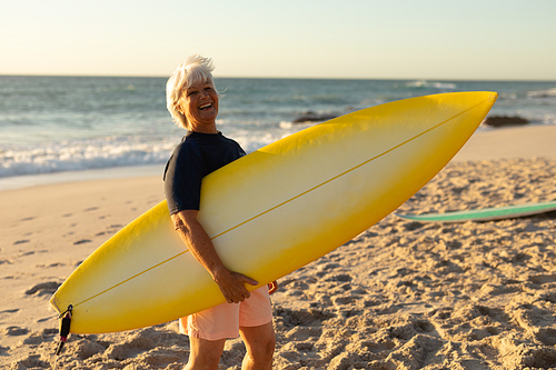 Side view of a senior Caucasian woman at the beach at sunset, standing on the sand holding a surfboard under her arm, looking to camera and smiling, with the sea in the bnackground