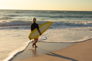 Side view of a senior Caucasian woman at the beach at sunset, walking on the sand carrying a surfboard under her arm, with the sea in the background