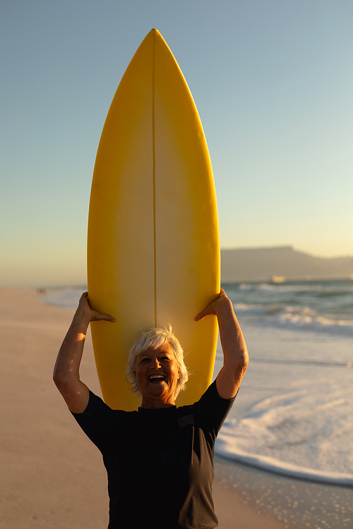 Front view close up of a senior Caucasian woman at the beach at sunset, standing on the sand holding a surfboard on her head, looking to camera and laughing