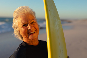 Front view close up of a senior Caucasian woman at the beach at sunset, standing on the sand holding a surfboard, looking to camera and laughing