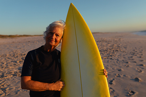 Front view of a senior Caucasian woman at the beach at sunset, standing on the sand holding a surfboard, looking to camera and smiling