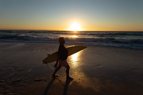 Side view silhouette of a senior Caucasian woman at the beach at sunset, walking on the sand towards the sea carrying a surfboard