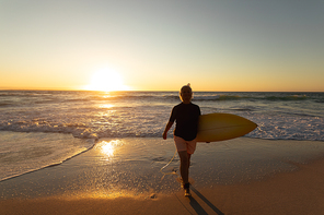 Front view of a senior Caucasian woman at the beach at sunset, walking on the sand towards the camera carrying a surfboard under her arm