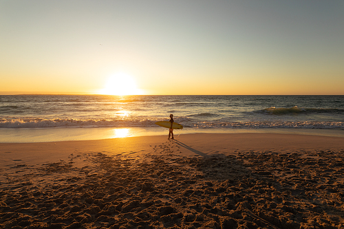 Distant side view of a senior Caucasian woman at the beach at sunset, walking on the sand by the sea carrying a surfboard