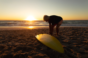 Side view of a senior Caucasian woman at the beach at sunset, standing on the sand by the sea attaching a surfboard to her leg rope