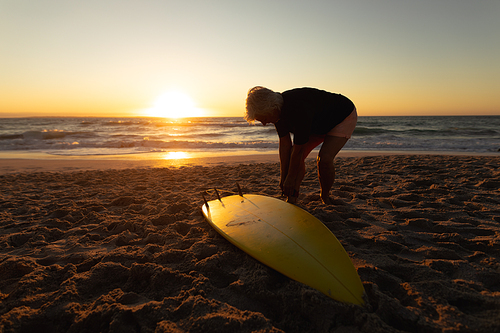 Side view of a senior Caucasian woman at the beach at sunset, standing on the sand by the sea attaching a surfboard to her leg rope