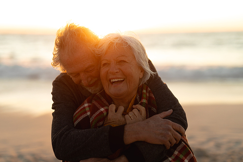 Front view close up of a senior Cacuasian couple standing on the beach at sunset smiling and embracing, with the sea in the background