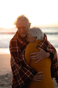 Front view close up of a senior Cacuasian couple standing on the beach at sunset with a blanket over their shoulders, smiling and embracing, with the sea in the background