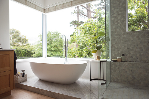 Modern bathroom interior showing free standing bath with large windows showing green trees and plants outside, with white bathtub and silver water tap, grey shower and a table with a plant.