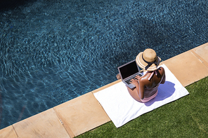 Mixed race woman spending time at home, relaxing and sunbathing by the pool, using a laptop. Self isolating and social distancing in quarantine lockdown during coronavirus covid 19 epidemic.