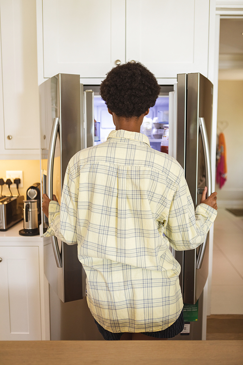 Mixed race woman spending time at home, standing in her kitchen and looking into a refrigerator. Self isolating and social distancing in quarantine lockdown during coronavirus covid 19 epidemic.