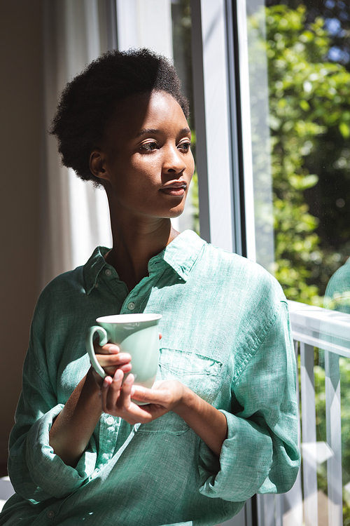 Mixed race woman spending time at home, holding a green mug and looking out the window. Self isolating and social distancing in quarantine lockdown during coronavirus covid 19 epidemic.