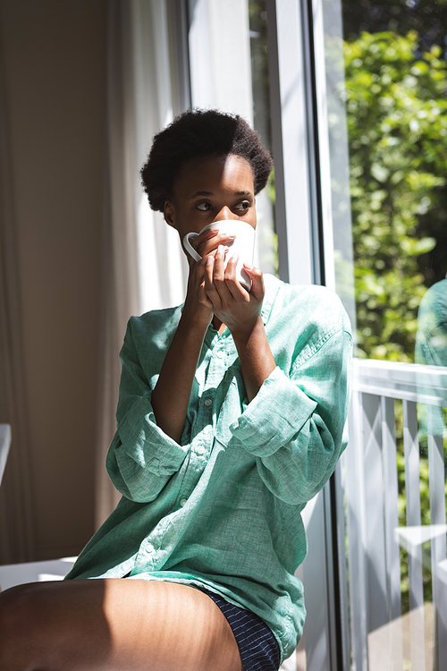 Mixed race woman spending time at home, holding a green mug and looking out the window. Self isolating and social distancing in quarantine lockdown during coronavirus covid 19 epidemic.