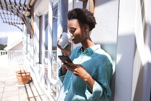 Mixed race woman spending time at home, standing at her terrace, holding a mug and using smartphone. Self isolating and social distancing in quarantine lockdown during coronavirus covid 19 epidemic.