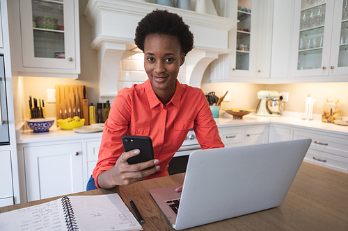 Mixed race woman spending time at home, sitting in her kitchen and using her laptop and smartphone. Self isolating and social distancing in quarantine lockdown during coronavirus covid 19 epidemic.