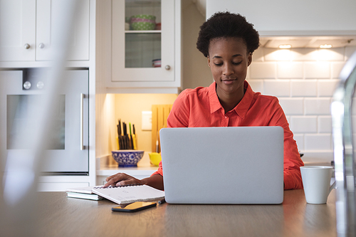 Mixed race woman spending time at home, sitting in her kitchen and using her laptop computer. Self isolating and social distancing in quarantine lockdown during coronavirus covid 19 epidemic.