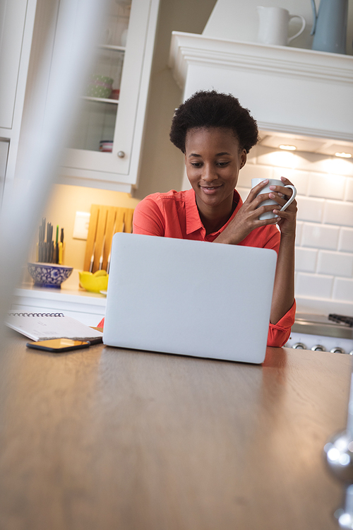 Mixed race woman spending time at home, sitting in her kitchen and using her laptop computer. Self isolating and social distancing in quarantine lockdown during coronavirus covid 19 epidemic.