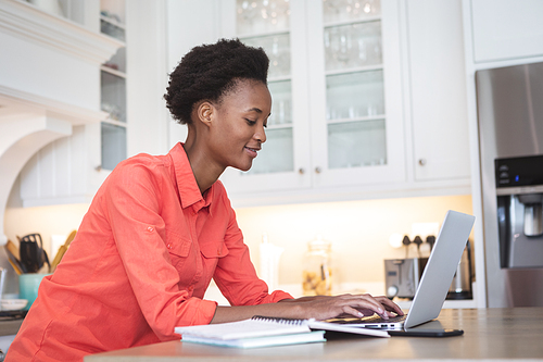 Mixed race woman spending time at home, sitting in her kitchen and using her laptop computer. Self isolating and social distancing in quarantine lockdown during coronavirus covid 19 epidemic.