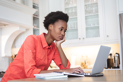 Mixed race woman spending time at home, sitting in her kitchen and using her laptop computer. Self isolating and social distancing in quarantine lockdown during coronavirus covid 19 epidemic.