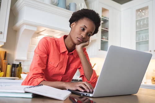 Mixed race woman spending time at home, sitting in her kitchen and using her laptop computer. Self isolating and social distancing in quarantine lockdown during coronavirus covid 19 epidemic.