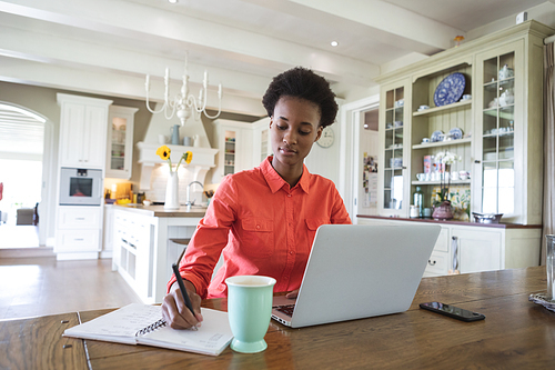 Mixed race woman spending time at home, sitting in her kitchen and using her laptop computer. Self isolating and social distancing in quarantine lockdown during coronavirus covid 19 epidemic.