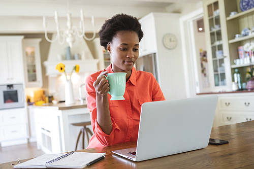 Mixed race woman spending time at home, sitting in her kitchen and using her laptop computer. Self isolating and social distancing in quarantine lockdown during coronavirus covid 19 epidemic.