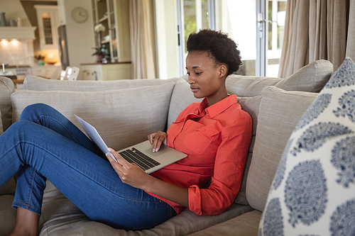 Mixed race woman spending time at home, sitting on couch in her in living room and using laptop. Self isolating and social distancing in quarantine lockdown during coronavirus covid 19 epidemic.