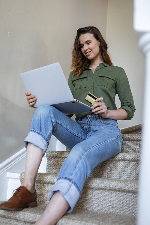 Caucasian woman spending time at home, sitting on stairs, using her laptop and credit card. Lifestyle at home isolating, social distancing in quarantine lockdown during coronavirus covid 19 pandemic.