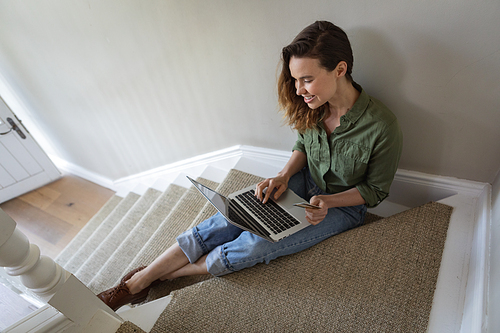 Caucasian woman spending time at home, sitting on stairs, using her laptop and credit card. Lifestyle at home isolating, social distancing in quarantine lockdown during coronavirus covid 19 pandemic.