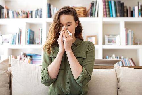 A Caucasian woman spending time at home, sitting on sofa, blowing her nose. Lifestyle at home isolating, social distancing in quarantine lockdown during coronavirus covid 19 pandemic.