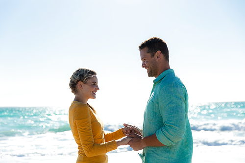 Front view of a Caucasian couple standing on the beach with blue sky and sea in the background, holding hands and smiling at each other