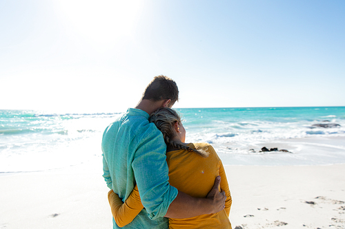 Rear view of a Caucasian couple standing on the beach with blue sky and sea in the background, embracing and looking away