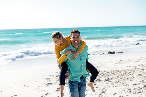 Front view of a Caucasian couple standing on the beach with blue sky and sea in the background, piggybacking and smiling