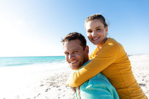 Side view of a Caucasian couple standing on the beach with blue sky and sea in the background, piggybacking and smiling to camera