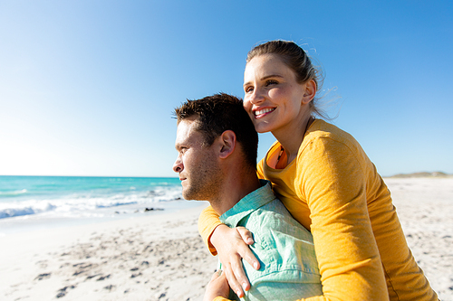 Side view of a Caucasian couple standing on the beach with blue sky and sea in the background, piggybacking, smiling and looking away