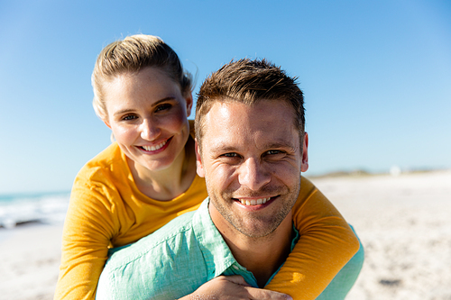 Portrait close up of a Caucasian couple standing on the beach with blue sky and sea in the background, piggybacking and smiling to camera