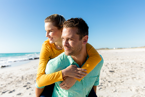 Front view of a Caucasian couple standing on the beach with blue sky and sea in the background, piggybacking and looking away